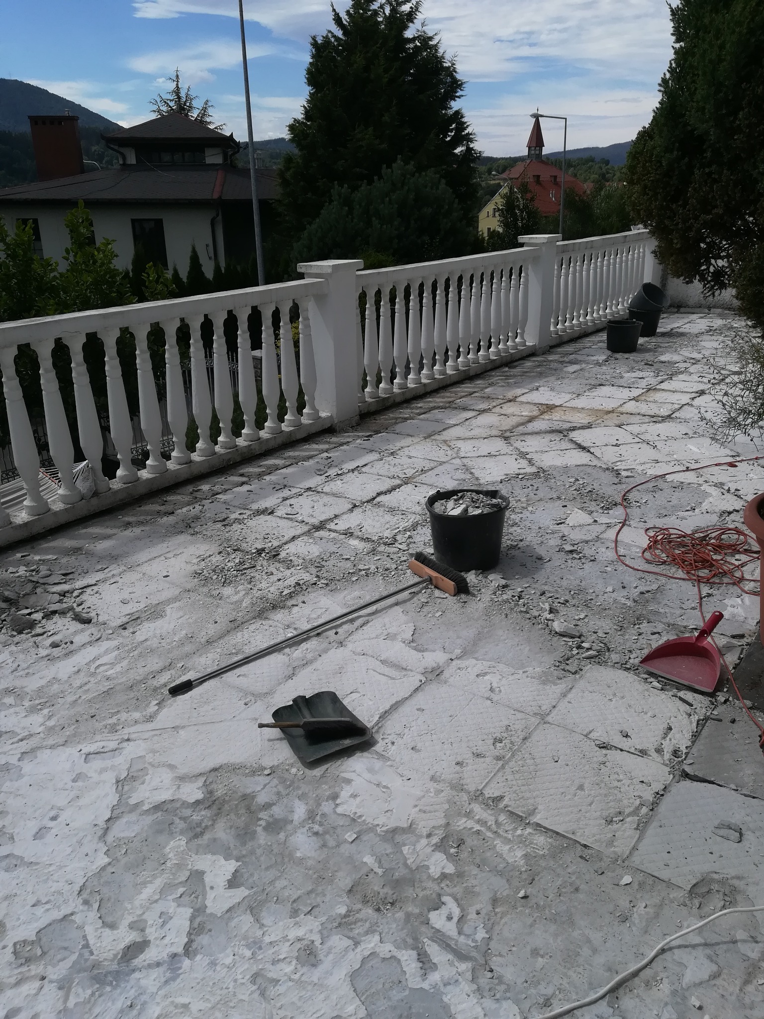 Taras w trakcie demontażu płytek, widoczne narzędzia: łopata, miotła, wiadro z gruzem, czerwony kabel, w tle biała balustrada i budynki.