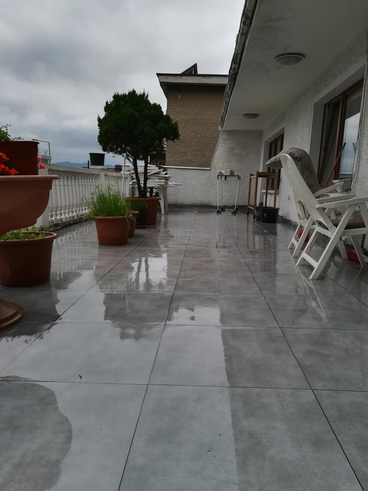 Błyszczący, szary taras po deszczu z rzędem donic z roślinami, białymi krzesłami i widokiem na góry w tle pod pochmurnym niebem.