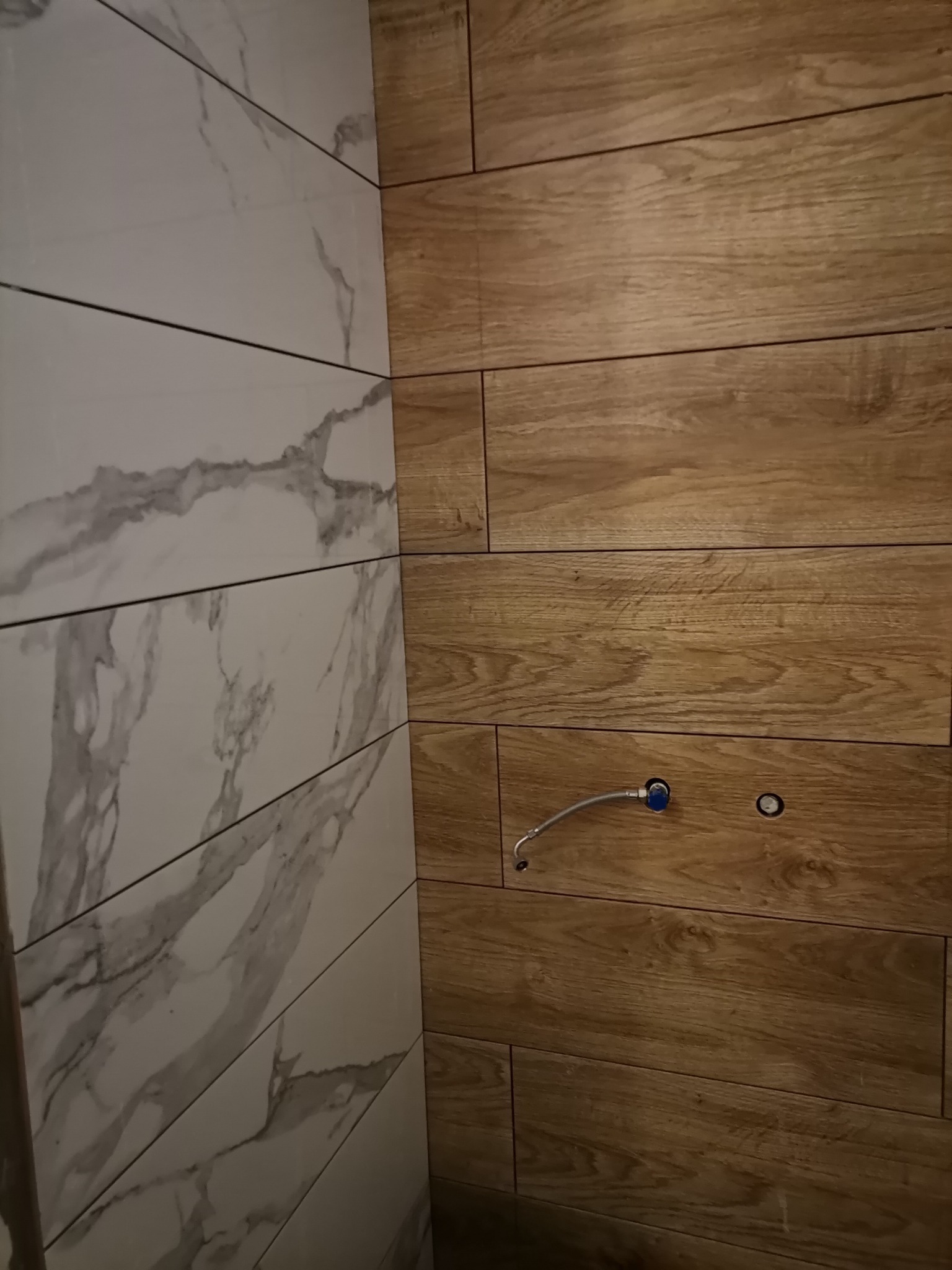 Ściana wyłożona płytkami imitującymi drewno i marmur, widoczne przyłącza wodne i otwór, wykonane w poziomie, z bliska.