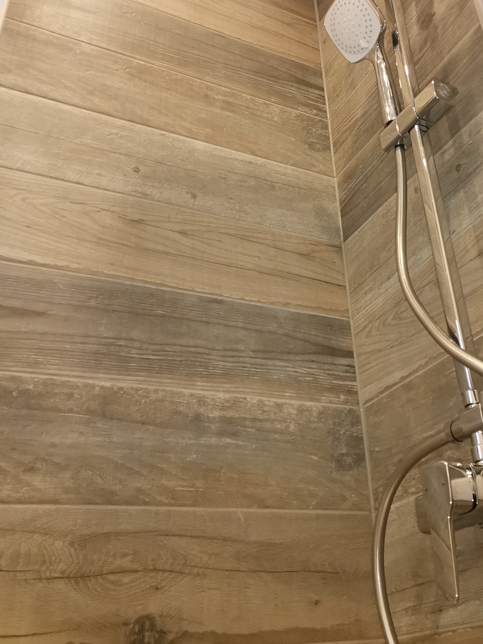 Wykończona ściana kabiny prysznicowej z płytek imitujących drewno i chromowana bateria z deszczownicą.