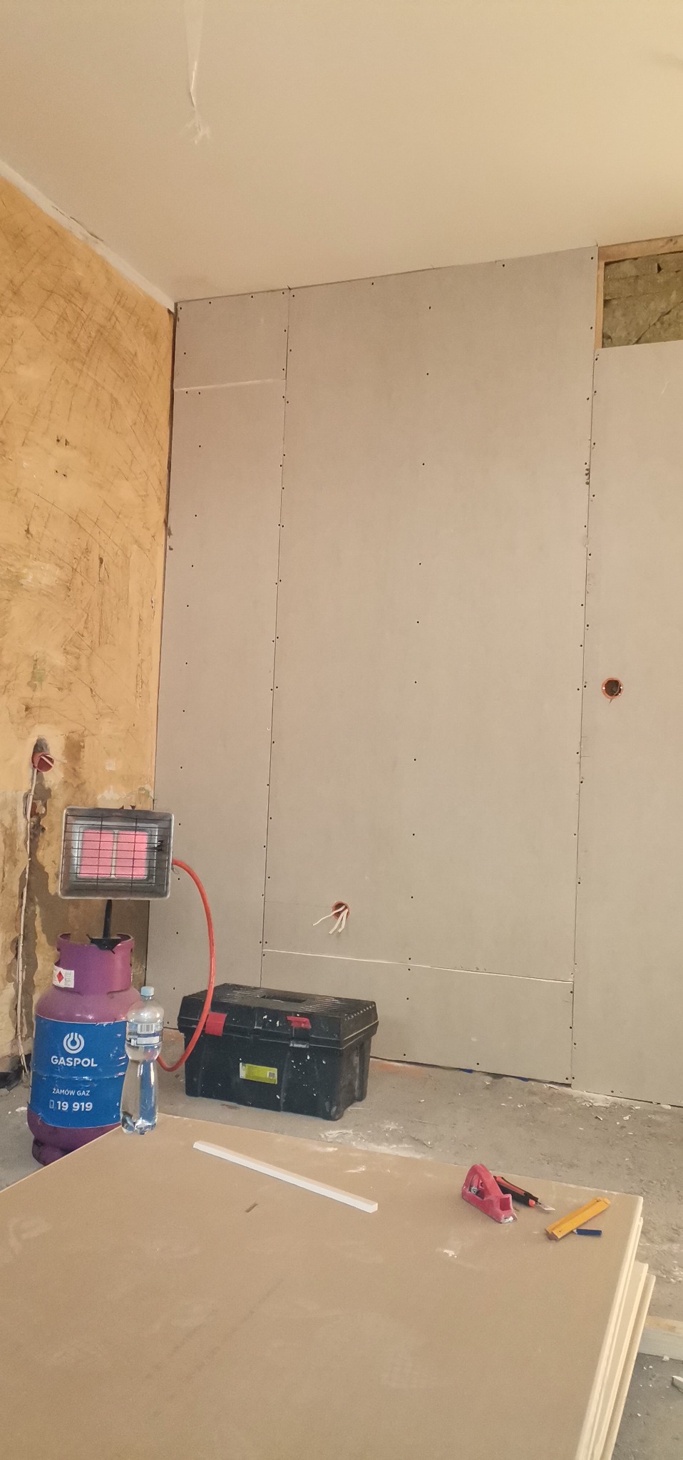 Wnętrze w trakcie montażu płyt kartonowo-gipsowych, widoczna butla gazowa z grzejnikiem, skrzynka z narzędziami i stos płyt. Ściana częściowo przygotowana, z widocznymi otworami na gniazdka.
