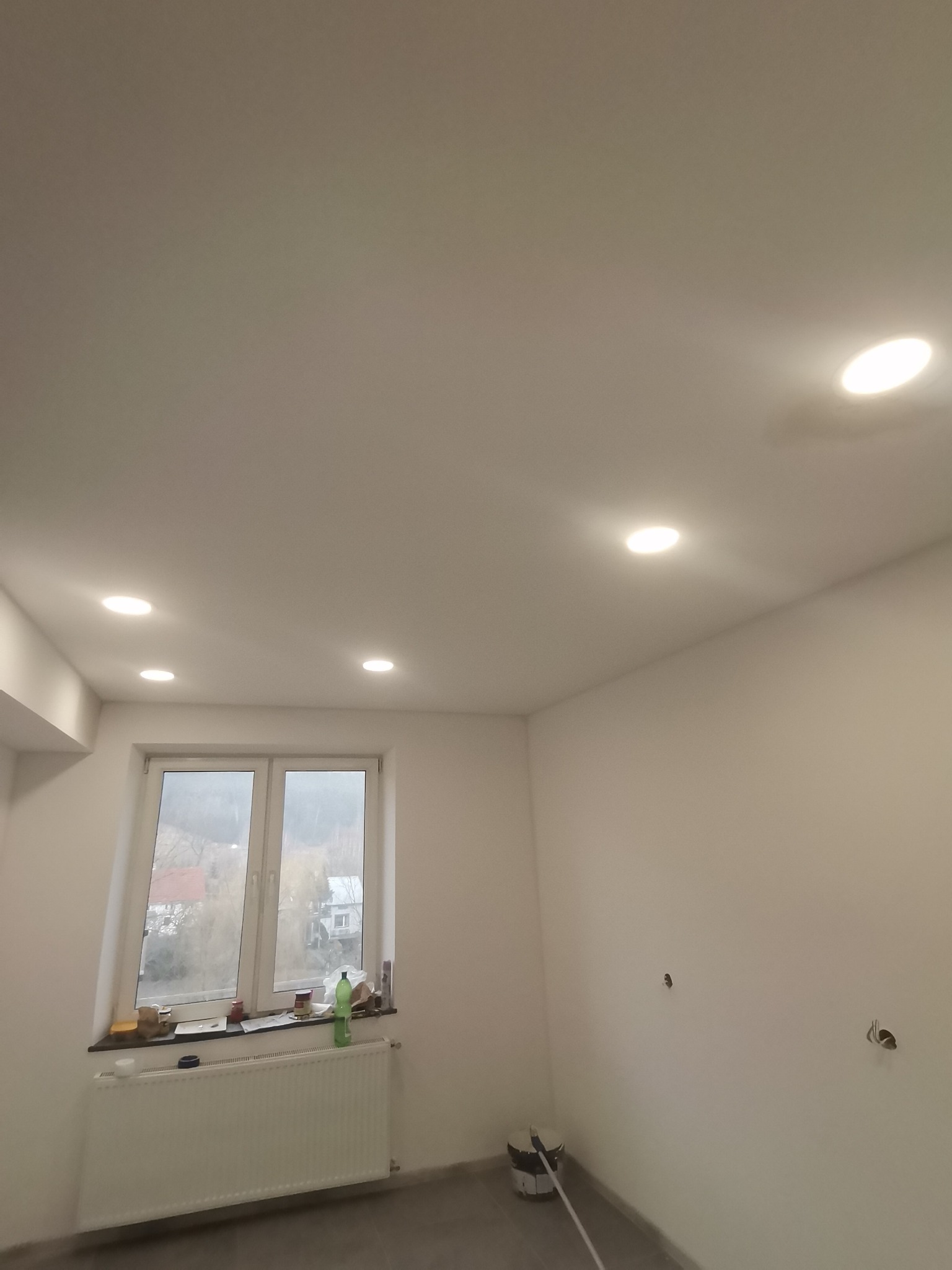 Wnętrze świeżo pomalowanego pokoju z trzema okrągłymi lampami halogenowymi w suficie, widok na okno z parapetem zastawionym różnymi przedmiotami, grzejnik pod oknem, ślady po gniazdkach na ścianie...