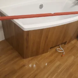 Biała wanna z obudową imitującą drewno, poziomica spoczywająca na jej krawędzi, podłoga z paneli w odcieniach brązu, widoczny kabel od urządzenia.