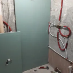 Montaż płyt kartonowo-gipsowych w łazience, widoczne instalacje hydrauliczne, narzędzia pracy: wiertarka, młotek, szczypce.