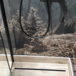 Kabina prysznicowa z czarną armaturą i odpływem liniowym na tle ściany z motywem leśnym.