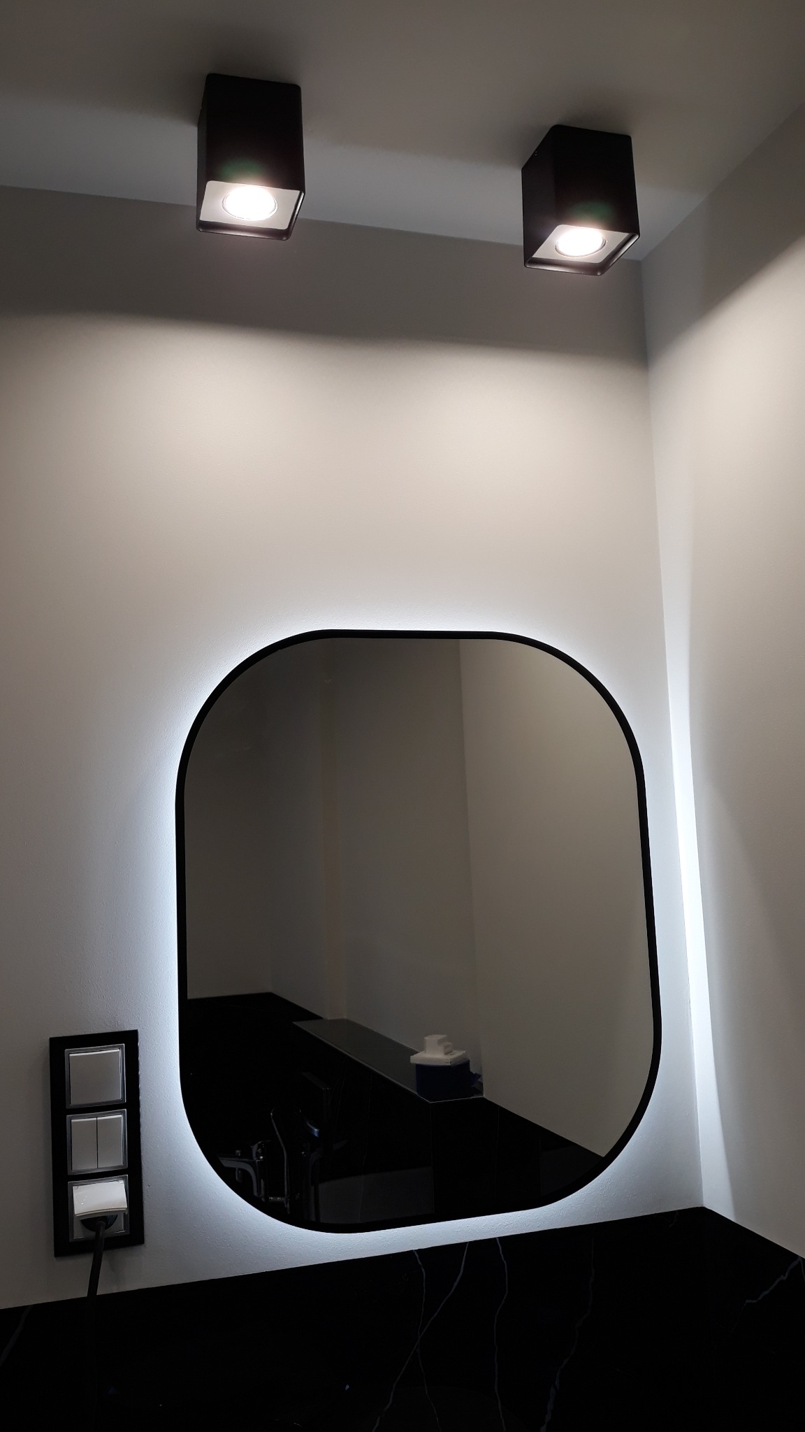 Nowoczesna łazienka z podświetlanym lustrem o zaokrąglonym kształcie, czarnym blatem imitującym marmur i minimalistycznymi czarnymi lampami sufitowymi, z widocznym panelem włączników światła.