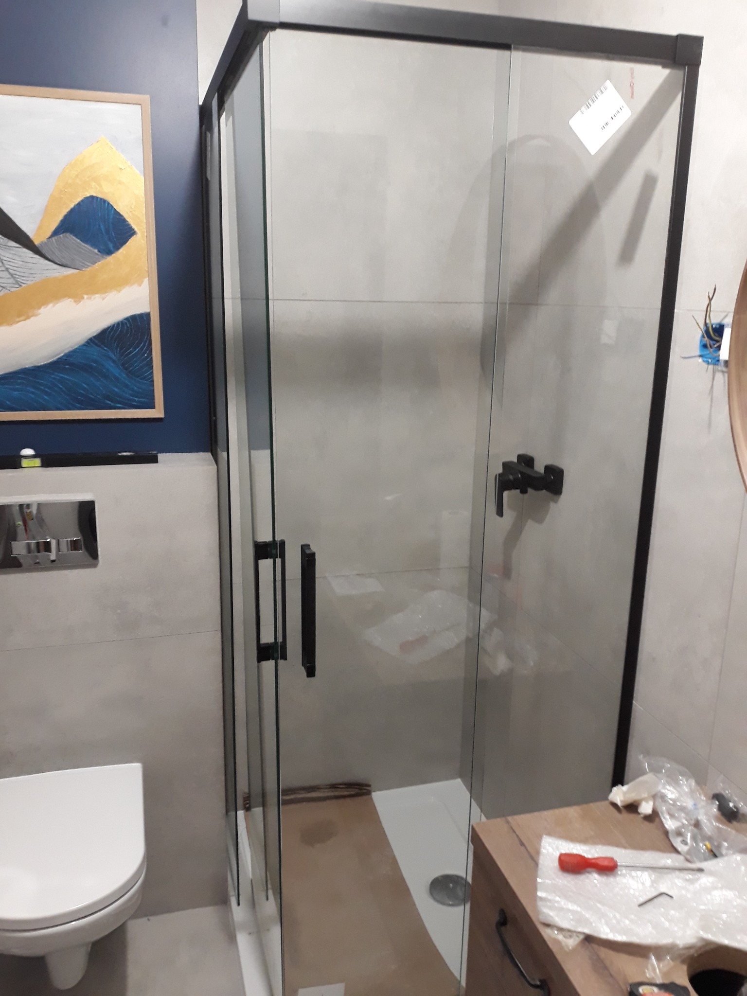 Nowoczesna kabina prysznicowa z czarnymi profilami i armaturą, zamontowana w łazience z szarymi płytkami i wiszącą toaletą, z obrazem na ścianie w tle i narzędziami na szafce.