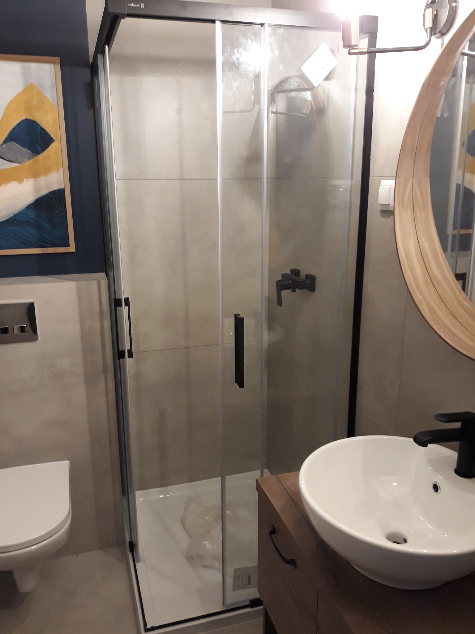 Nowoczesna łazienka z kabiną prysznicową z czarnymi detalami, okrągłym lustrem w drewnianej ramie i umywalką nablatową na drewnianej szafce. W tle widoczna toaleta i obraz na ścianie.