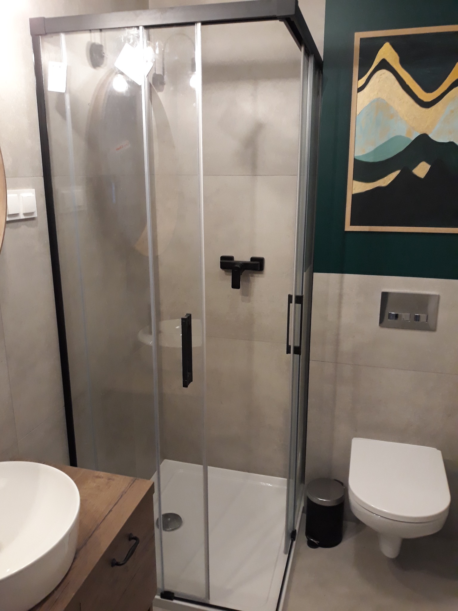 Nowoczesna kabina prysznicowa z czarnymi profilami i armaturą, widok na umywalkę nablatową i toaletę w łazience z akcentem ciemnej zieleni oraz obrazem w stylu abstrakcyjnym.