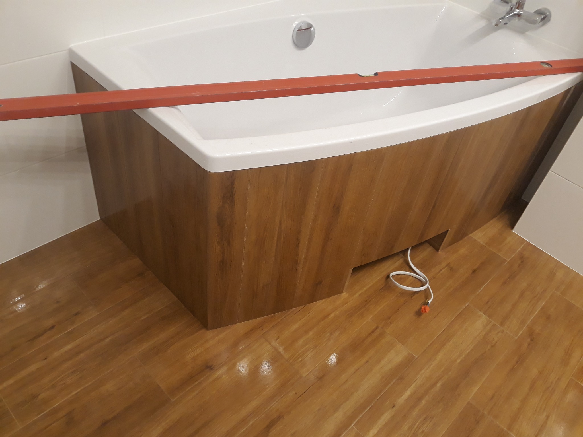 Biała wanna z obudową imitującą drewno, poziomica spoczywająca na jej krawędzi, podłoga z paneli w odcieniach brązu, widoczny kabel od urządzenia.