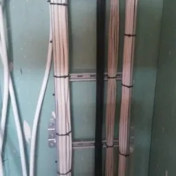 Wiązki białych kabli elektrycznych przymocowane czarnymi opaskami do metalowej szyny montażowej na zielonej ścianie z płyt gipsowo-kartonowych w trakcie instalacji.