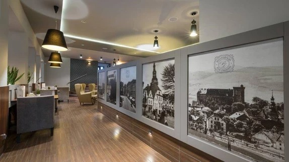 Wnętrze restauracji z drewnianą podłogą, szarymi ścianami i dużymi czarno-białymi fotografiami przedstawiającymi zabytkowe budynki i krajobrazy. Czarne, stożkowe lampy wiszą nad stolikami z szarymi...