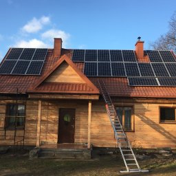 Solar - Ekspert s.c - Dom z nową instalacją paneli słonecznych na dachu z czerwonej dachówki, dwie drabiny opierają się o budynek, a obok stoi osoba w czarnym ubraniu i masce.