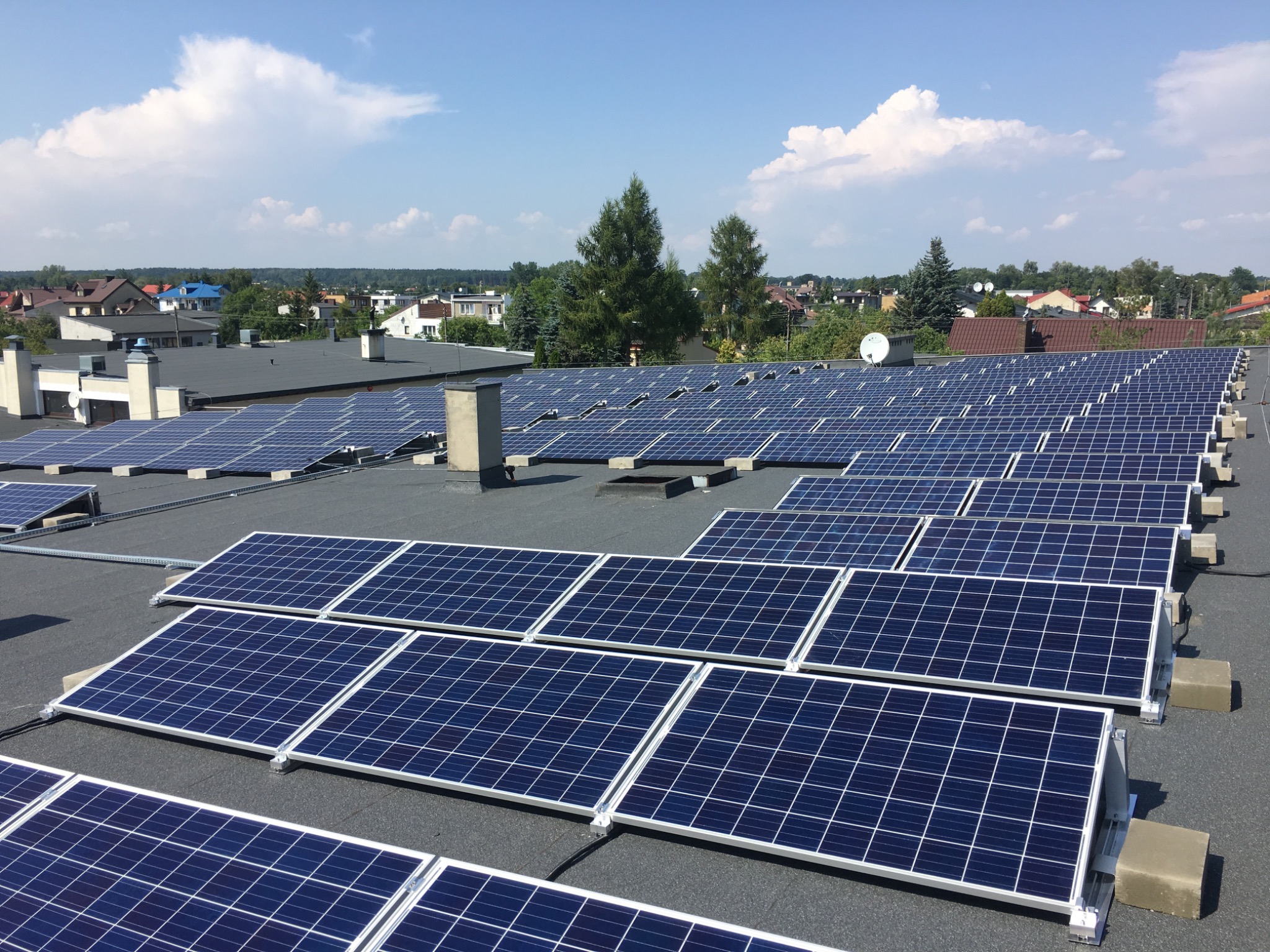 Instalacja paneli słonecznych na płaskim dachu budynku, widok z góry na tle zabudowy jednorodzinnej i błękitnego nieba z chmurami.