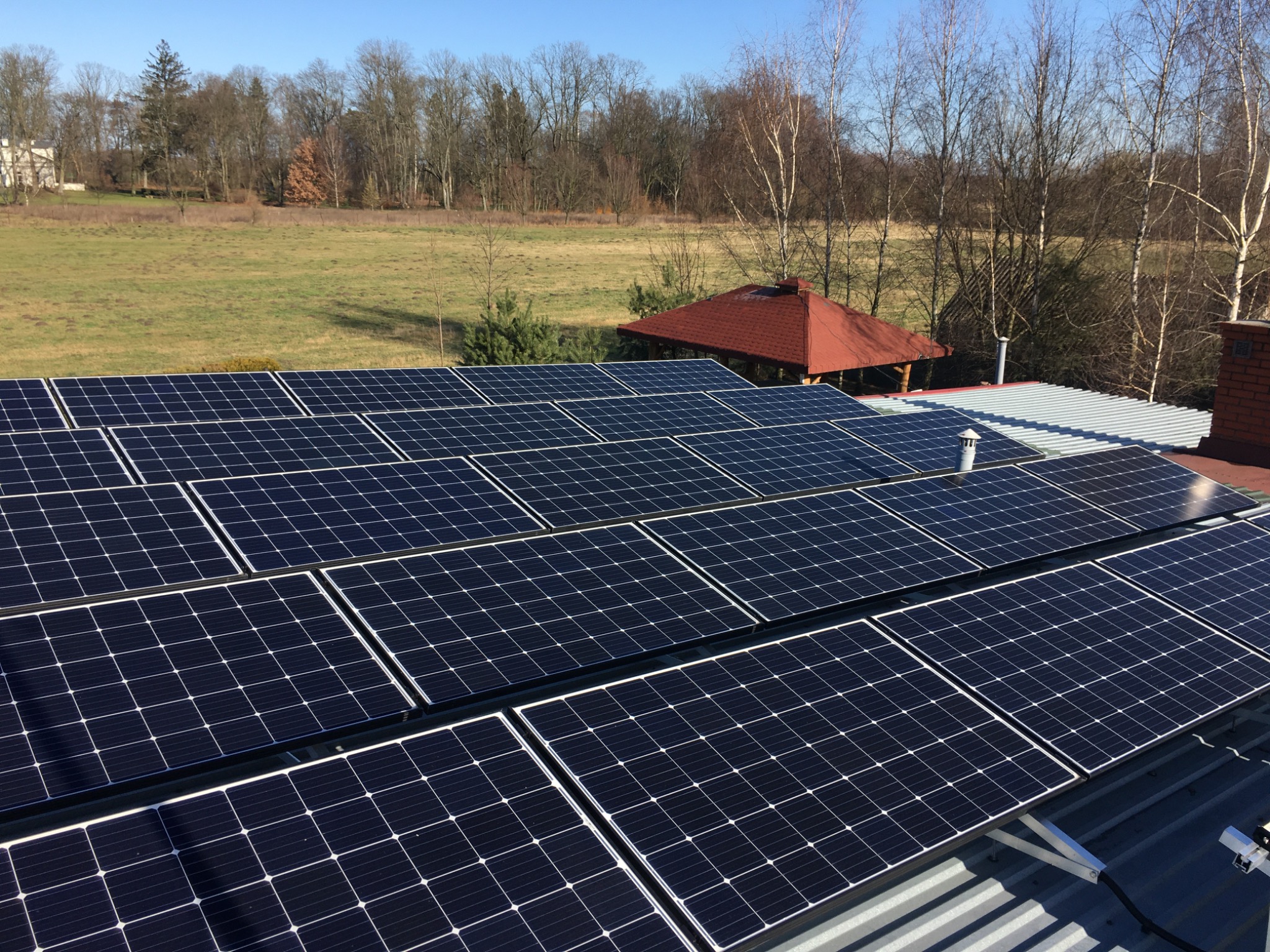 Rząd paneli słonecznych na metalowym dachu domu jednorodzinnego, w tle zielone pole, drzewa i altana z czerwonym dachem w słoneczny dzień.