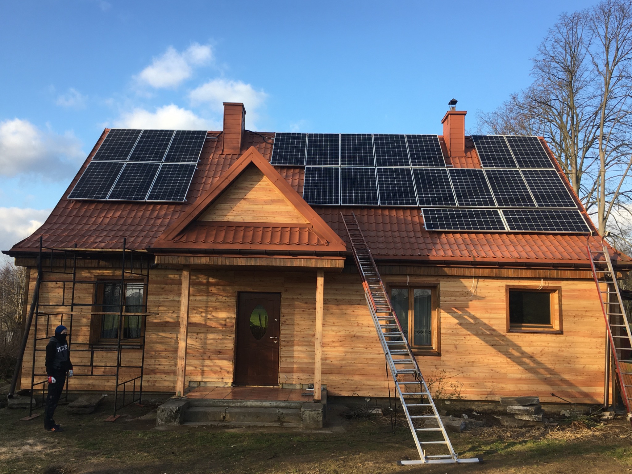 Dom z nową instalacją paneli słonecznych na dachu z czerwonej dachówki, dwie drabiny opierają się o budynek, a obok stoi osoba w czarnym ubraniu i masce.