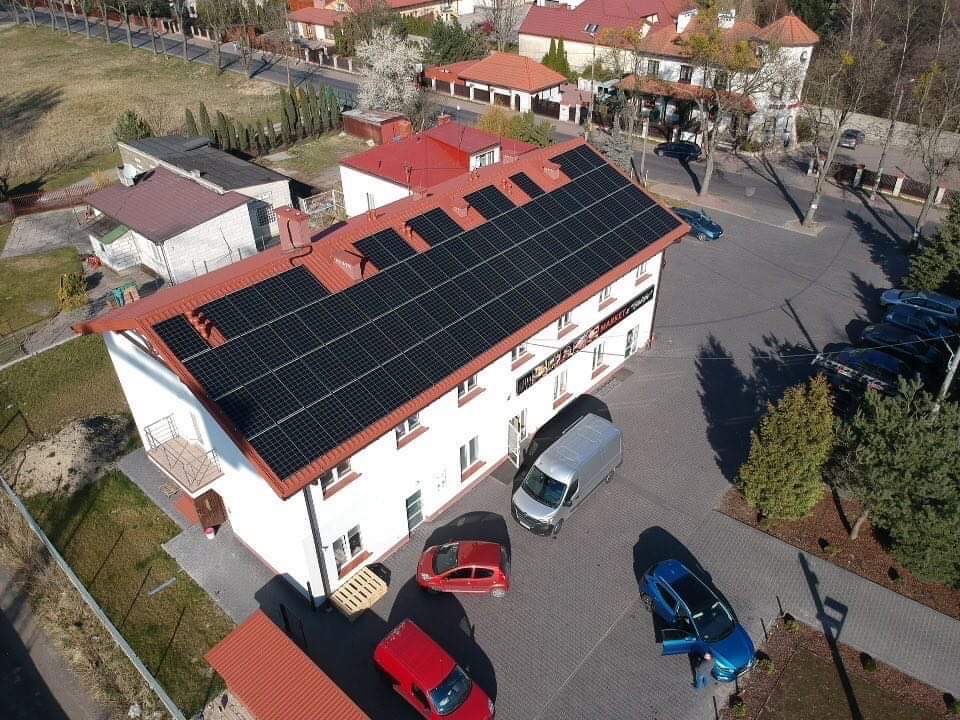 Widok z lotu ptaka na budynek z panelami słonecznymi na dachu, zaparkowane samochody i chodnik z kostki brukowej.