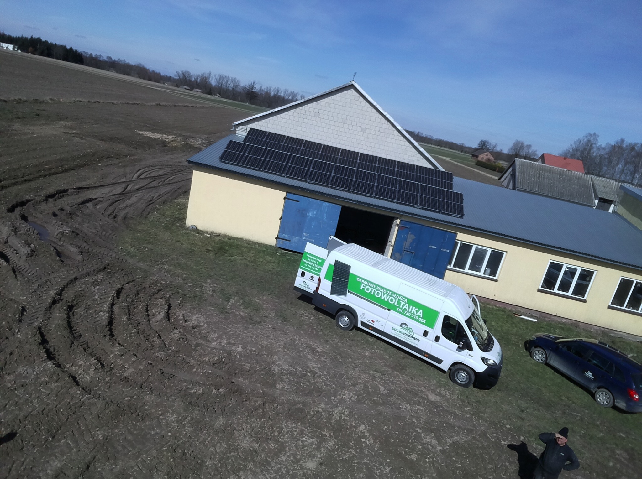 Instalacja fotowoltaiczna na dachu budynku gospodarczego z zaparkowanym obok samochodem firmowym z logo firmy fotowoltaicznej.