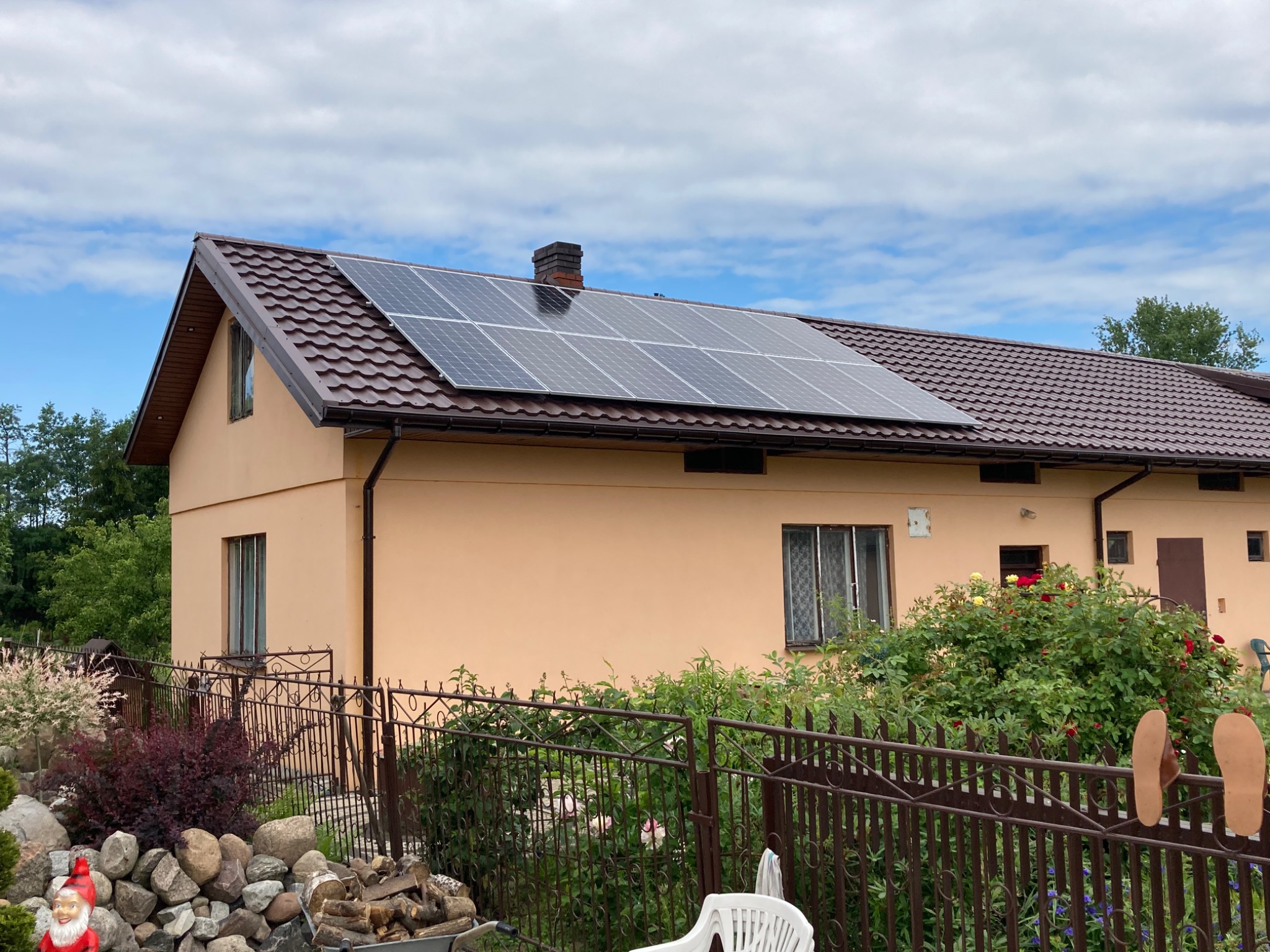 Dom jednorodzinny z brązowym dachem z dachówki i panelami słonecznymi na dachu, widok z ogrodu z ozdobnymi kamieniami i roślinami.