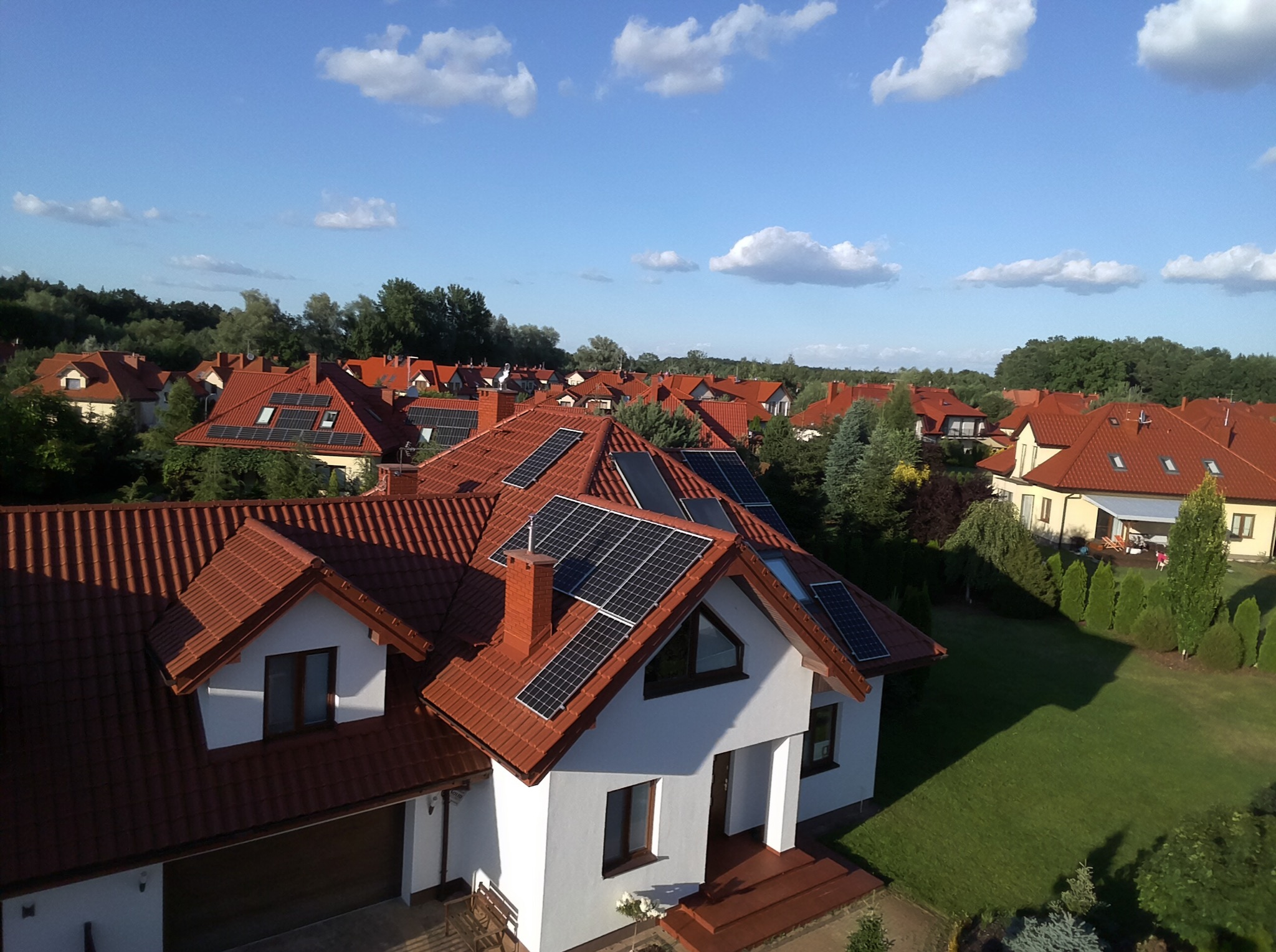 Widok z góry na osiedle domów jednorodzinnych z panelami słonecznymi na dachach, w tle drzewa i błękitne niebo z chmurami.