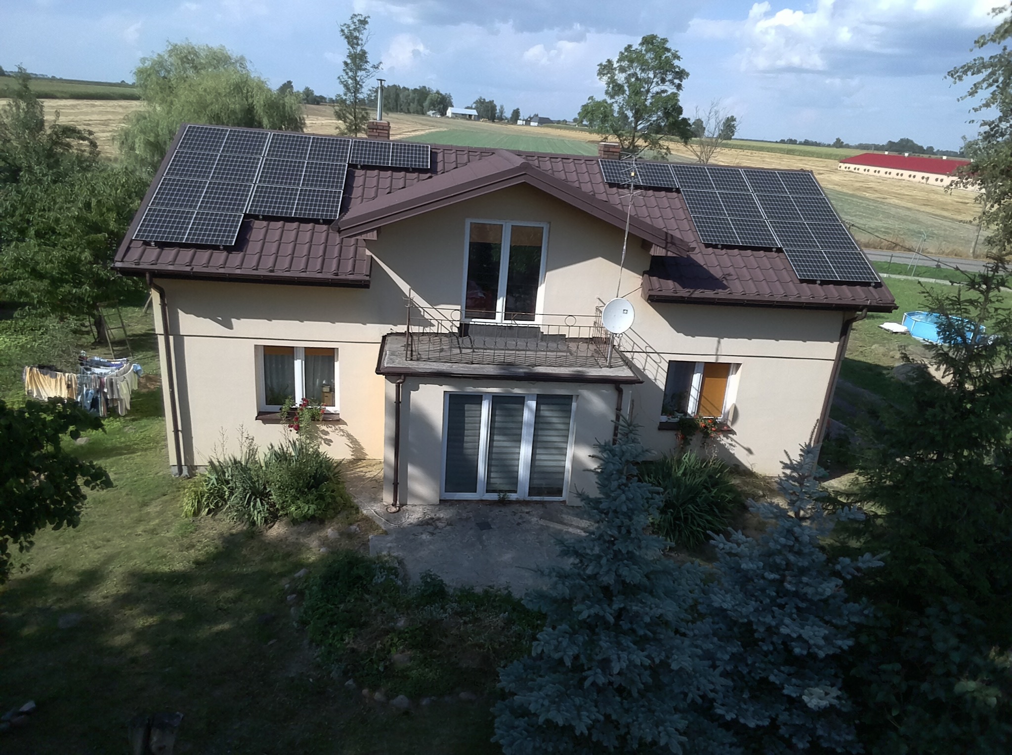 Dom jednorodzinny z brązowym dachem, na którym zamontowano panele słoneczne; widok z góry, w tle pola i budynki gospodarcze.
