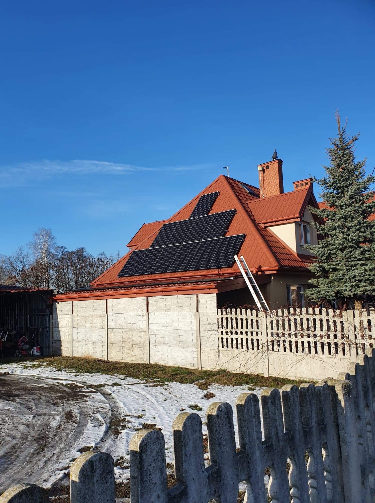 Dom jednorodzinny z czerwoną dachówką i panelami słonecznymi na dachu. Przystawiona drabina sugeruje trwające prace instalacyjne. Częściowo ośnieżone podwórko i betonowe ogrodzenie w tle.
