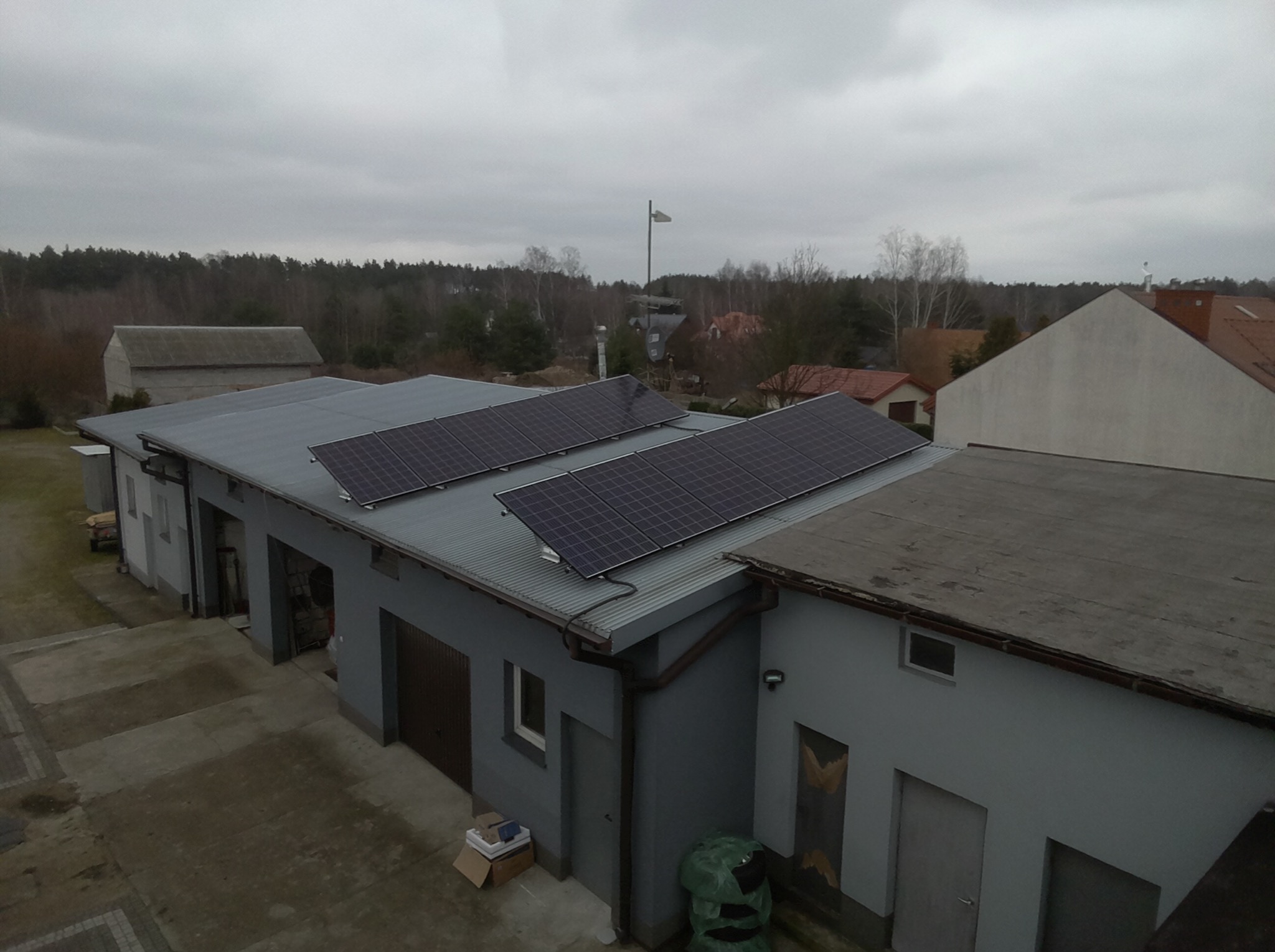 Widok z góry na szary budynek z trzema panelami fotowoltaicznymi na dachu z blachy falistej; w tle drzewa i pochmurne niebo.