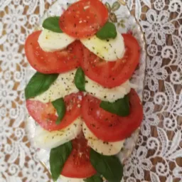 Caprese na ozdobnym talerzu z motywem kwiatowym na białym, koronkowym obrusie. Plasterki pomidorów i mozzarelli przełożone listkami bazylii, posypane przyprawami.