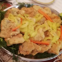 Srebrna patera z porcją smażonej ryby w panierce, udekorowanej zielonym koperkiem, liściem sałaty i paskami marynowanej papryki, na tle wzorzystego obrusa.