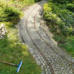 Zakrzywiony chodnik z kostki granitowej z czerwonymi wstawkami, częściowo zasypany piaskiem, z widoczną miotłą na trawie, w otoczeniu zieleni i krzewów.