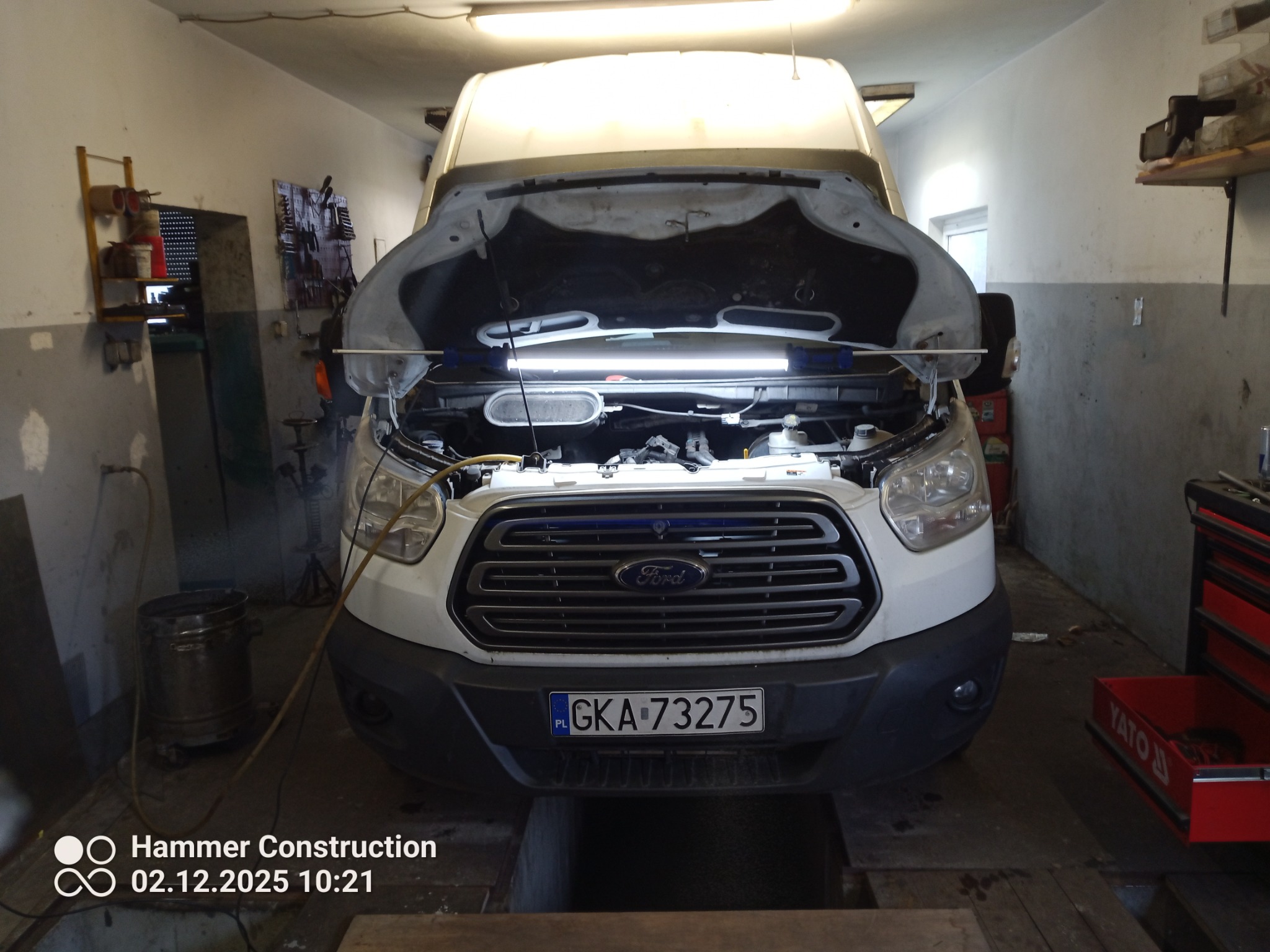 Biały Ford Transit z otwartą maską w warsztacie. Widoczna lampa oświetlająca silnik, narzędzia na ścianie i skrzynka Yato. Tablica rejestracyjna GKA 73275.