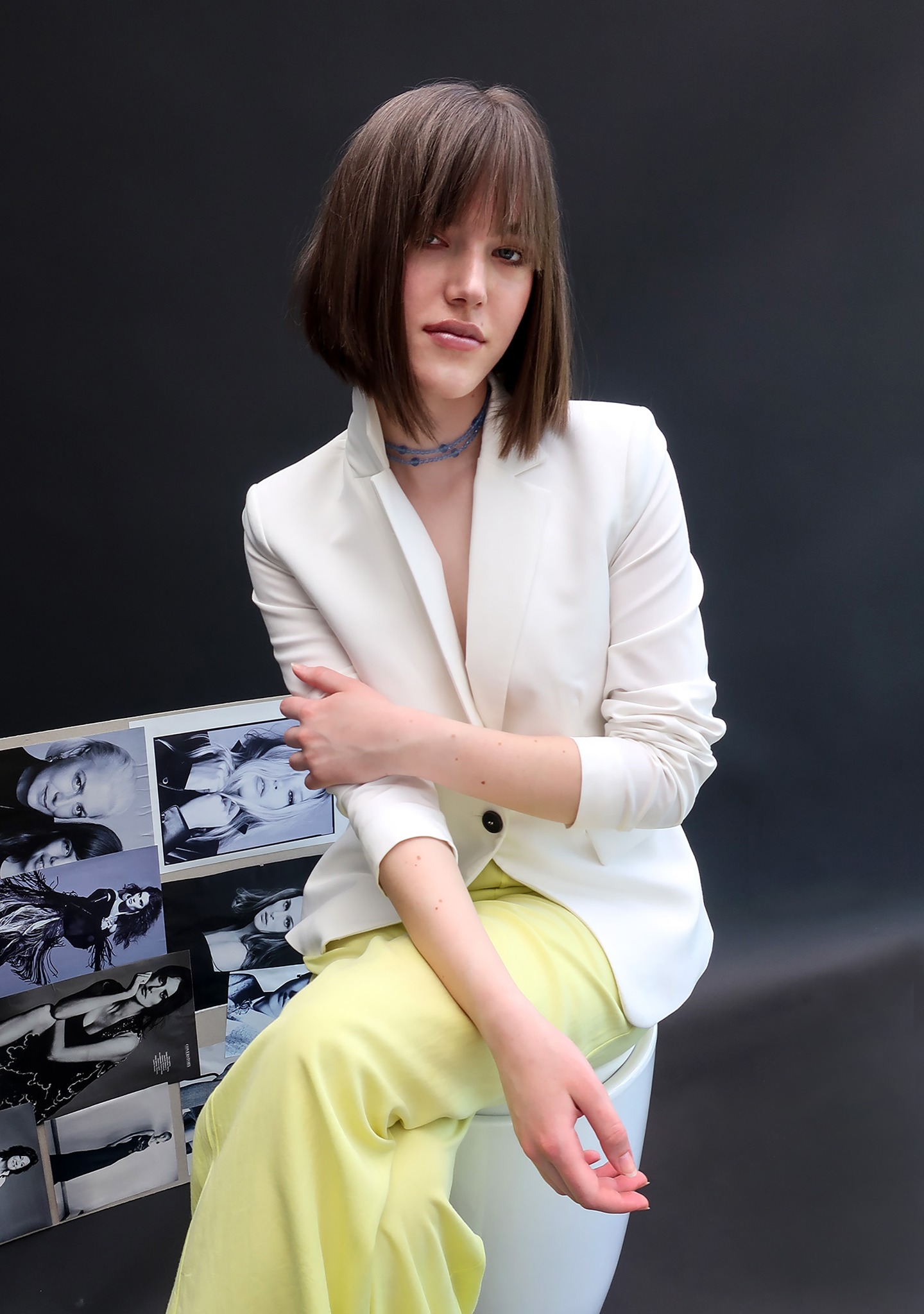 Portret młodej kobiety w białym żakiecie i żółtych spodniach, siedzącej na krześle obok kolażu czarno-białych zdjęć portretowych na ciemnym tle.
