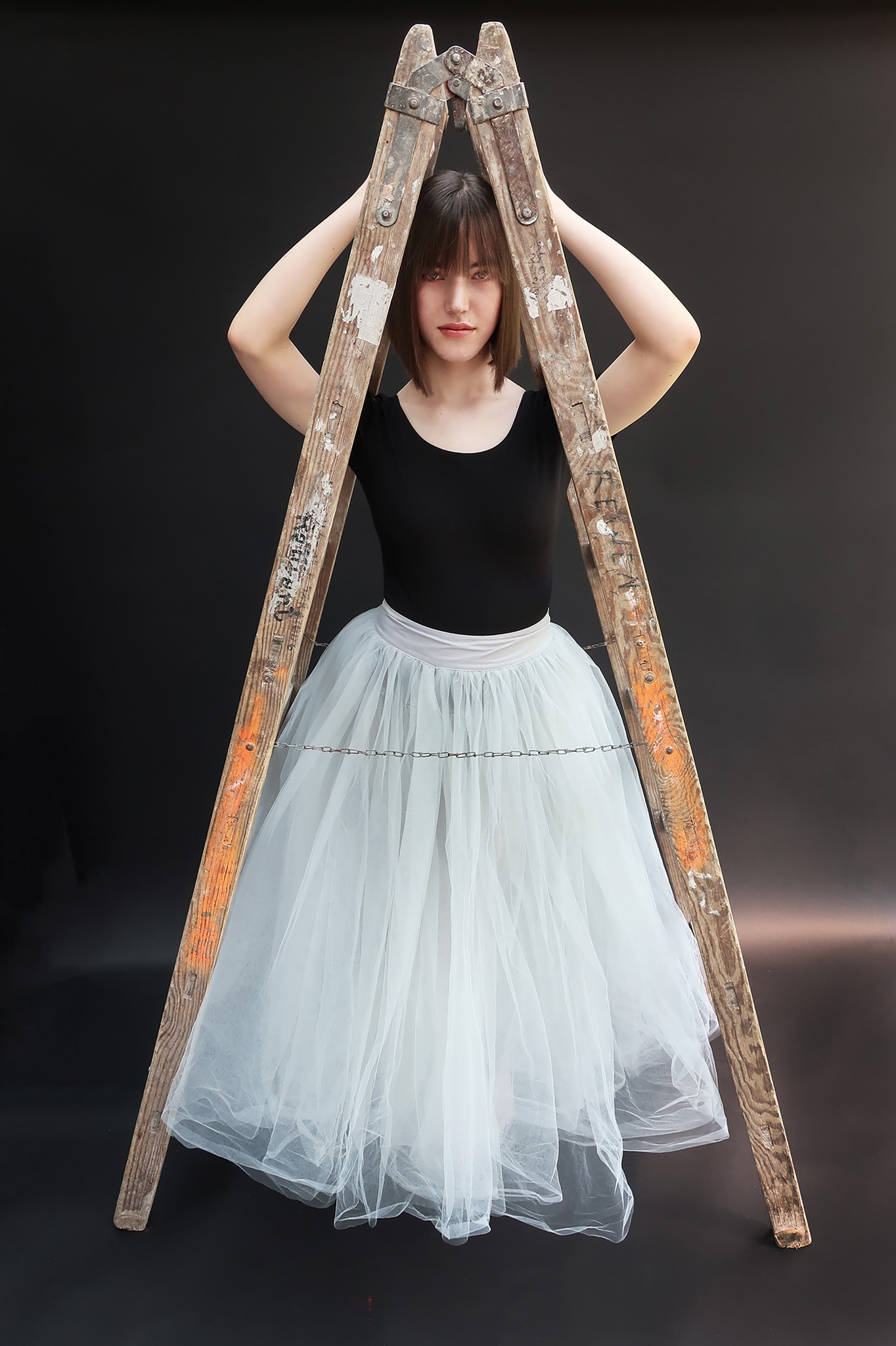 Portret młodej kobiety w tiulowej spódnicy, pozującej z drewnianą drabiną na czarnym tle.