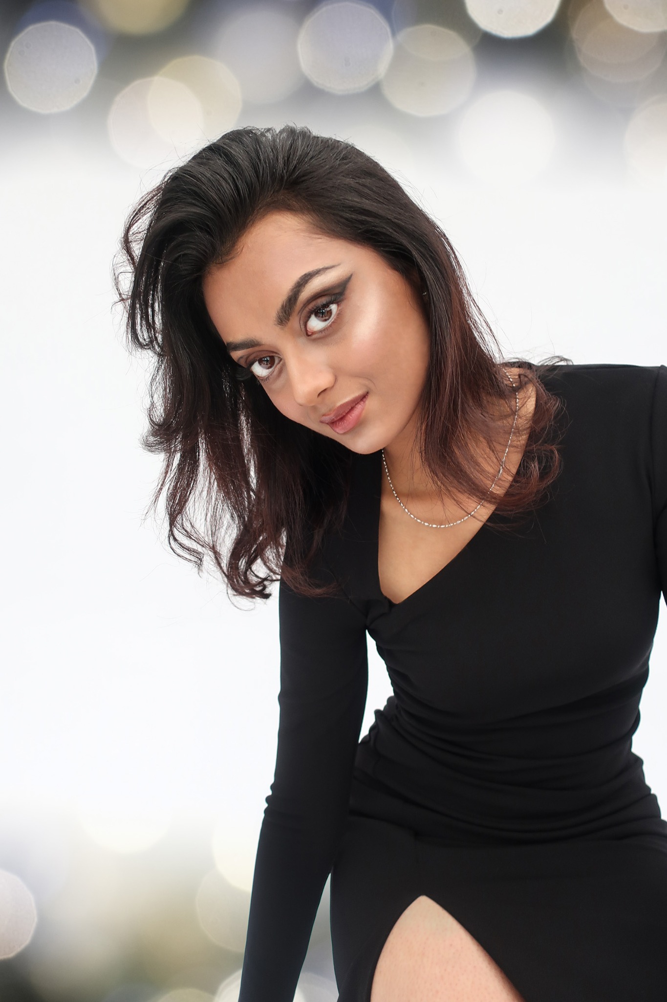 Portret kobiety w czarnej sukience, z mocnym makijażem oczu i srebrnym naszyjnikiem, pozującej na rozmytym, jasnym tle.