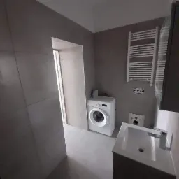 Wykończona łazienka w Gliwicach z szarymi płytkami, białą pralką, grzejnikiem drabinkowym i umywalką z szafką, widok z perspektywy