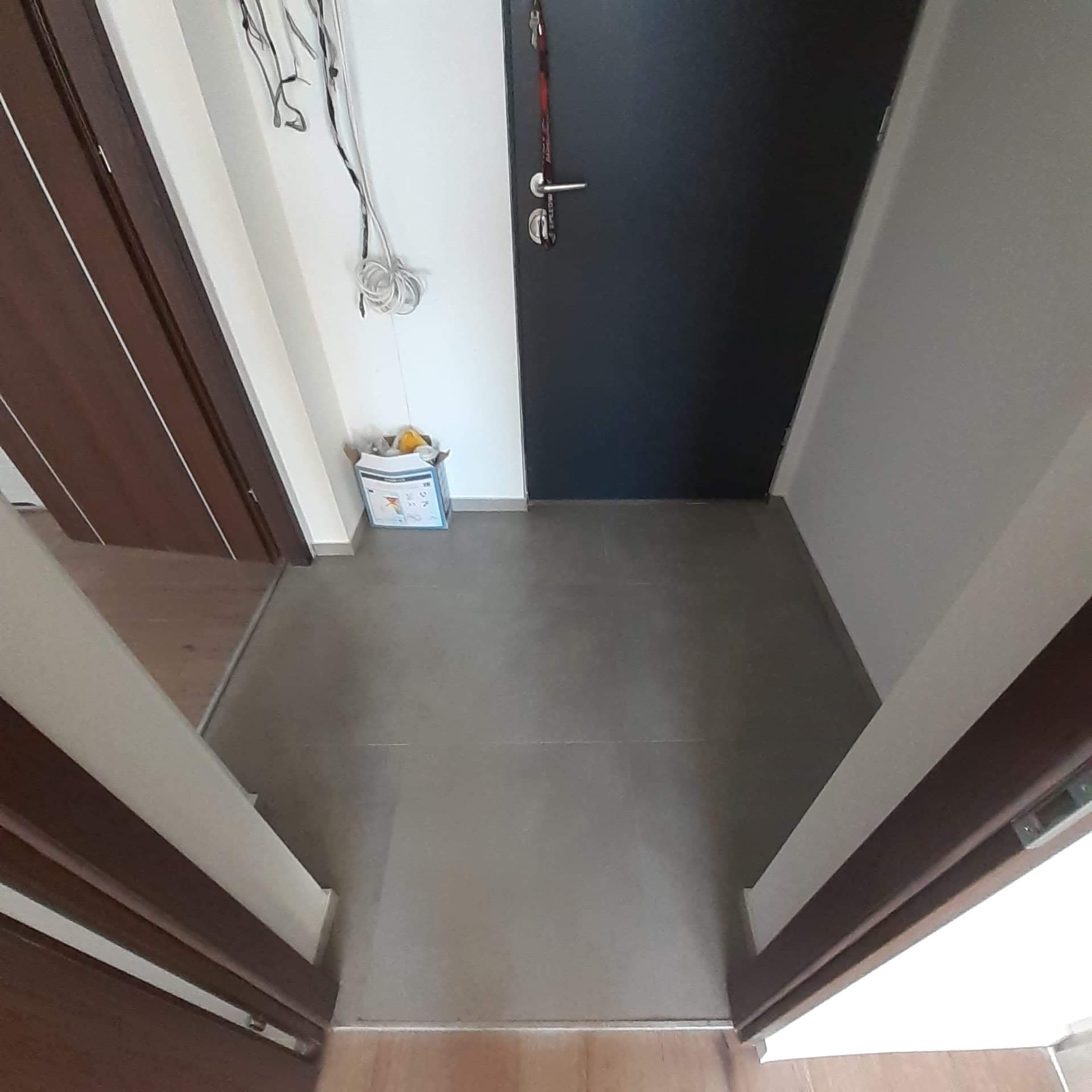 Wykończona podłoga w przedpokoju z szarych, dużych płytek, widoczna czarna futryna drzwi wejściowych oraz karton po płytkach przy ścianie.