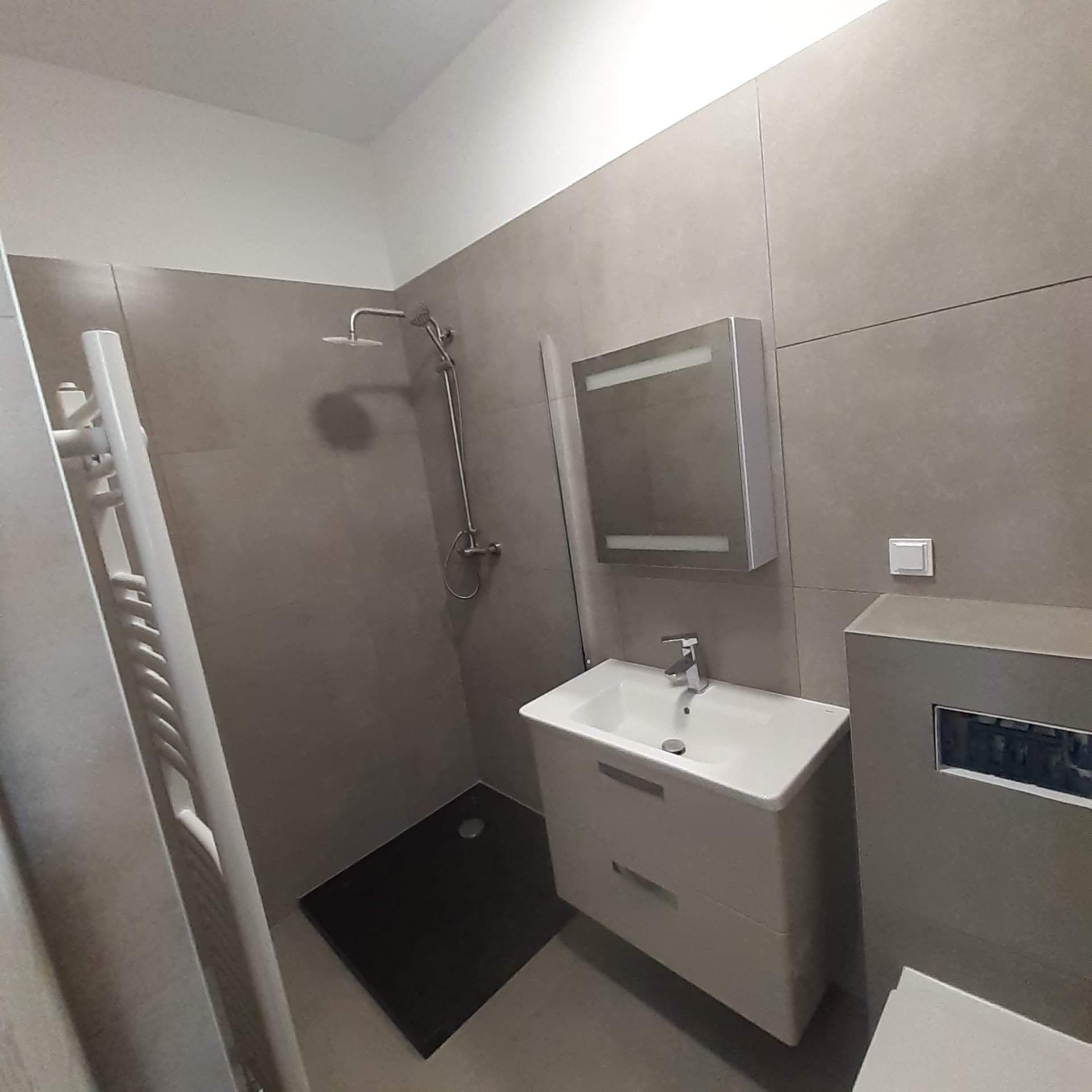 Wykończona łazienka z szarymi płytkami, kabiną prysznicową typu walk-in z czarnym brodzikiem, białą umywalką z szafką i grzejnikiem drabinkowym.