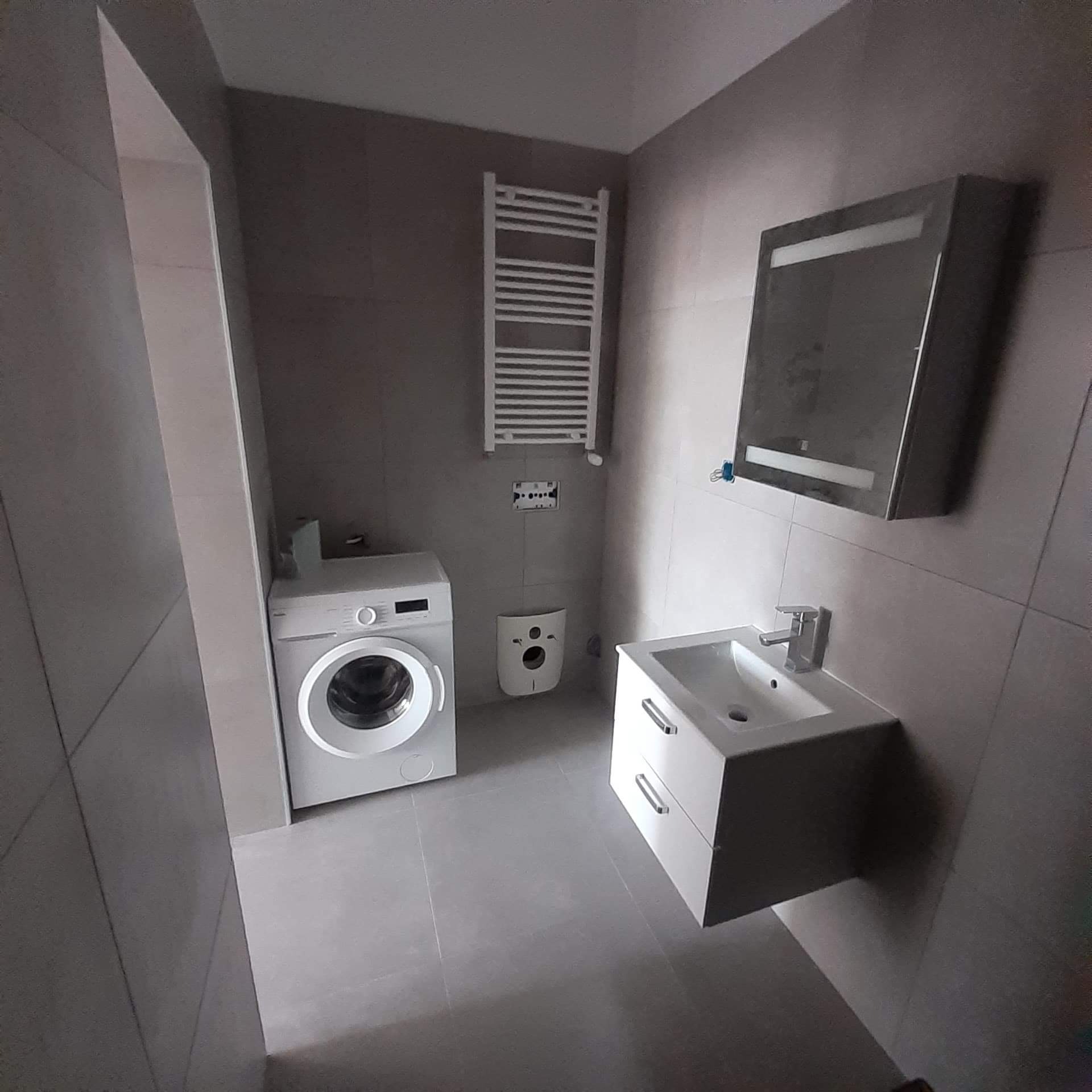 Wykończona łazienka w szarych płytkach, z białą pralką, grzejnikiem drabinkowym, umywalką z szafką i lustrem z oświetleniem LED.