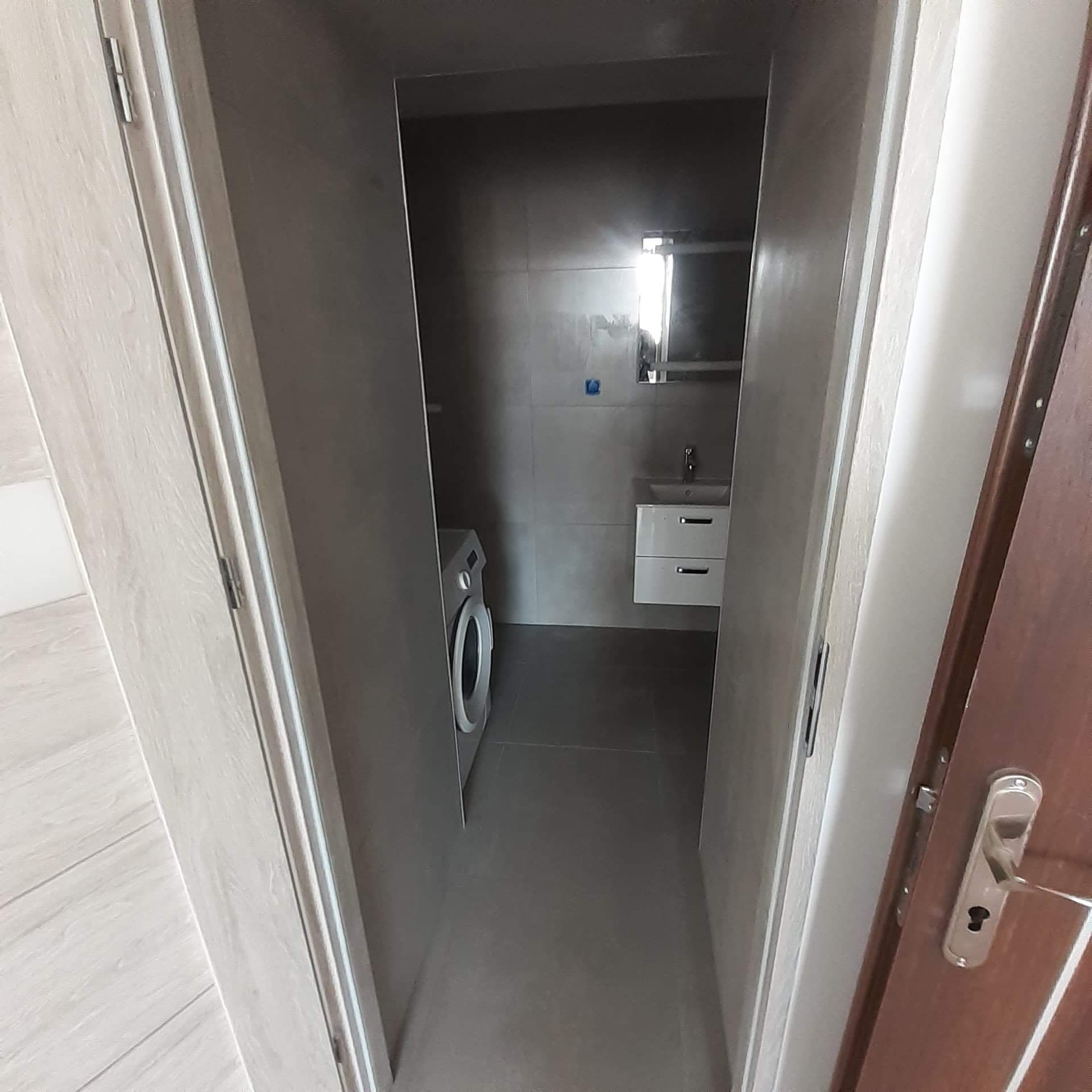 Wąskie wnętrze łazienki z szarymi płytkami na ścianach i podłodze, widok z korytarza przez otwarte drzwi, pralka po lewej stronie, umywalka z szafką i lustrem na końcu.