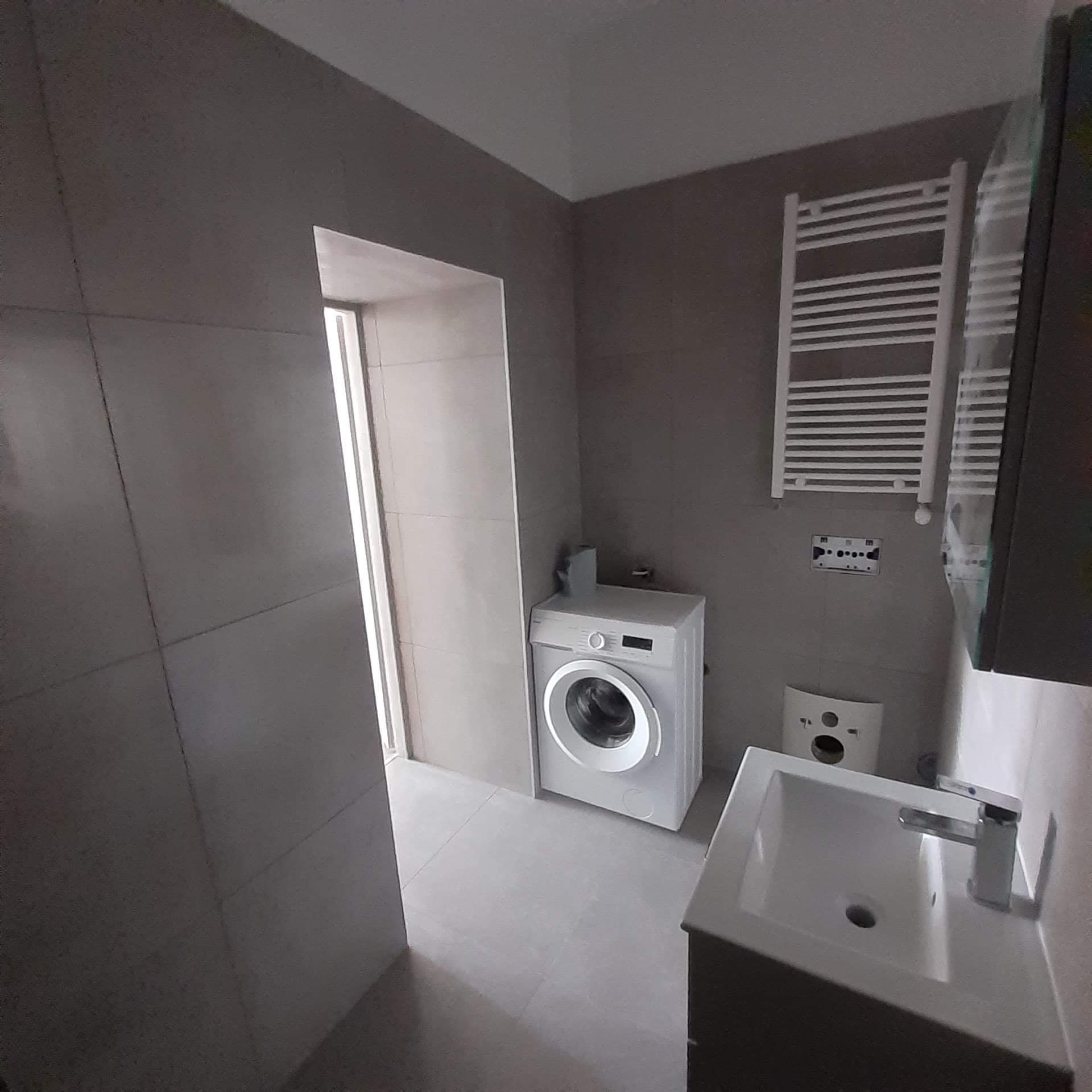 Wykończona łazienka w Gliwicach z szarymi płytkami, białą pralką, grzejnikiem drabinkowym i umywalką z szafką, widok z perspektywy