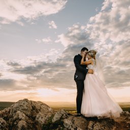 ARTIST MEDIA RAFAŁ GŁOGOWSKI MICHAŁ ZIÓŁKOWSKI s.c. - Para młoda, on w ciemnym garniturze, ona w sukni z koronkową górą i delikatnym różowym dołem, stoi na skalistym wzgórzu na tle dramatycznego, zachodzącego słońca i chmur.