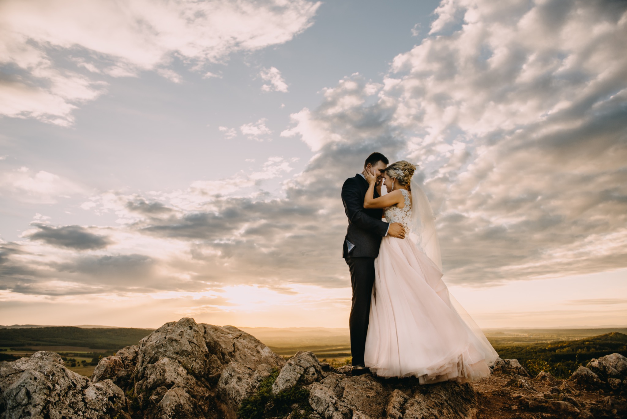 Para młoda, on w ciemnym garniturze, ona w sukni z koronkową górą i delikatnym różowym dołem, stoi na skalistym wzgórzu na tle dramatycznego, zachodzącego słońca i chmur.