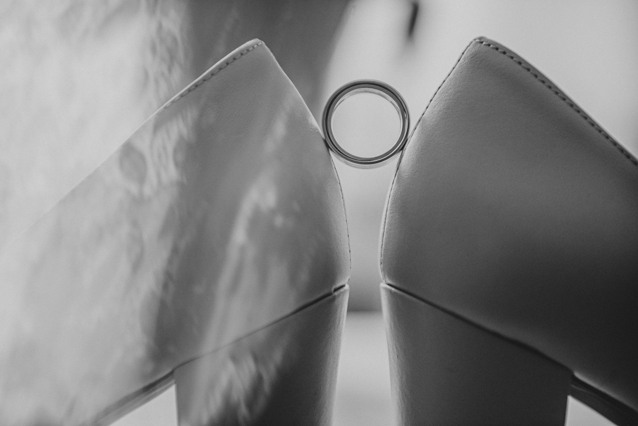 Czarno-białe zbliżenie dwóch czółenek damskich ustawionych obok siebie, z obrączką ślubną umieszczoną pomiędzy nimi, na tle delikatnej tkaniny.