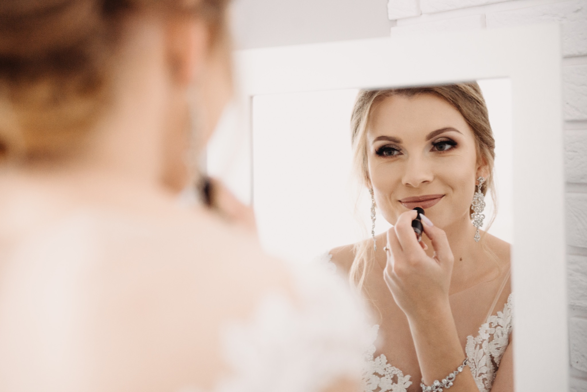 Panna młoda w sukni ślubnej, patrząc w lustro, aplikuje szminkę; widoczne duże kolczyki i bransoletka.