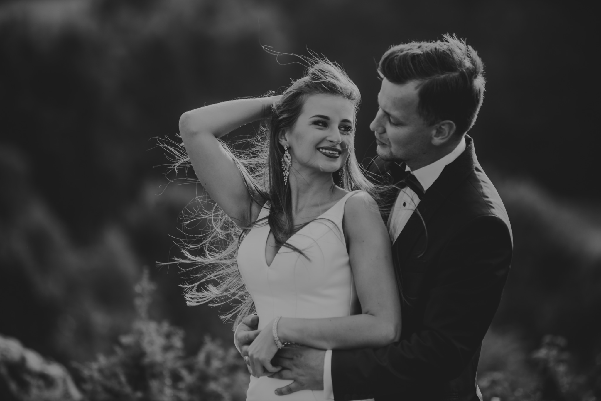 Czarno-białe zdjęcie uśmiechniętej panny młodej w białej sukni, z rozwianymi włosami, obejmowanej przez pana młodego w smokingu z muchą.