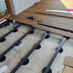 Montaż tarasu kompozytowego: widoczne legary na regulowanych wspornikach, częściowo ułożone deski, wkrętarka i nóż monterski obok