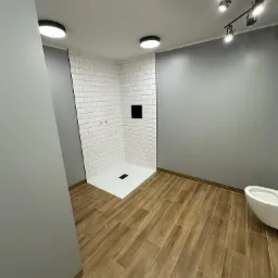 Wykończona łazienka w Białymstoku z kabiną prysznicową wyłożoną białymi, prostokątnymi płytkami, drewnopodobną podłogą i szarą ścianą, oświetlona punktowymi lampami.