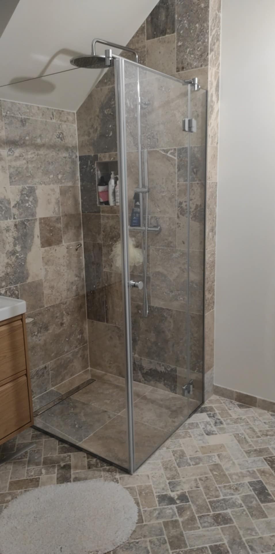 Nowoczesna kabina prysznicowa typu walk-in z deszczownicą, wykończona płytkami w odcieniach szarości i beżu, widoczna półka w ścianie z kosmetykami i dywanik na podłodze ułożonej w jodełkę.