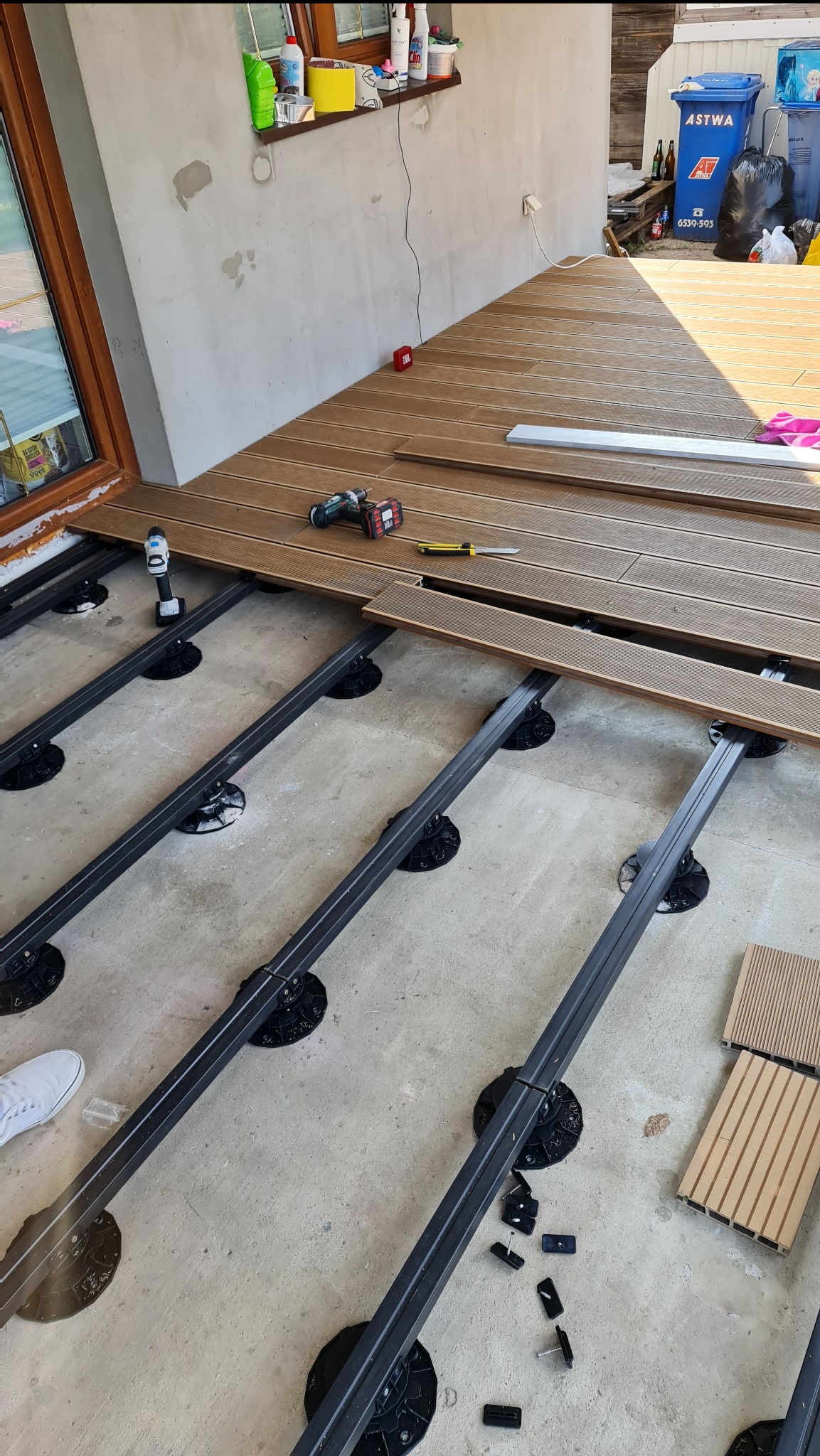 Montaż tarasu kompozytowego: widoczne legary na regulowanych wspornikach, częściowo ułożone deski, wkrętarka i nóż monterski obok