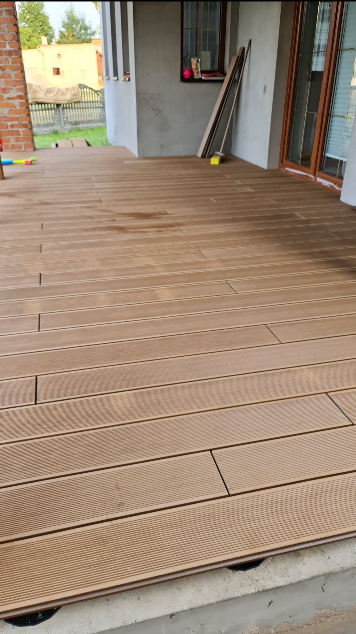 Nowo ułożony taras z desek kompozytowych o ciepłym, brązowym odcieniu, widoczne elementy mocujące przy betonowej wylewce, zarys budynku w tle.